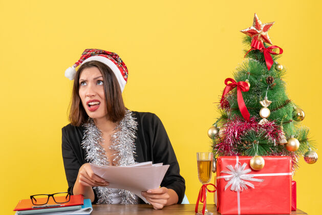 成人穿着西装 戴着圣诞老人帽和新年装饰品 手里拿着文件 坐在一张桌子旁 桌上放着圣诞树的商务女士震惊了文件漂亮年