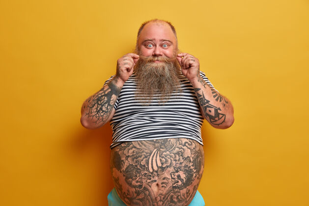 姿势滑稽的胡须男人的肖像卷曲的胡子 有纹身的胳膊和肚子 穿着小尺寸的条纹无袖t恤 有肥胖和超重的问题 隔离在黄色的墙上大触摸肥胖