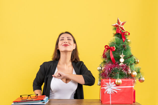 成功满意的年轻女子在黄色办公室装饰的圣诞树旁 穿着西装享受她的成功享受树套装