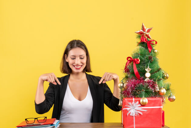 圣诞在黄色的办公室里 一个穿着西装的满意的年轻女人指着装饰好的圣诞树肖像套装黄色