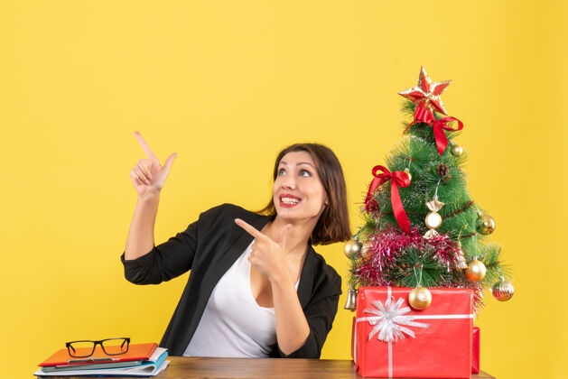 成人情绪激动的年轻女子指着坐在黄色办公室装饰圣诞树旁的桌子上的东西时尚坐着桌子
