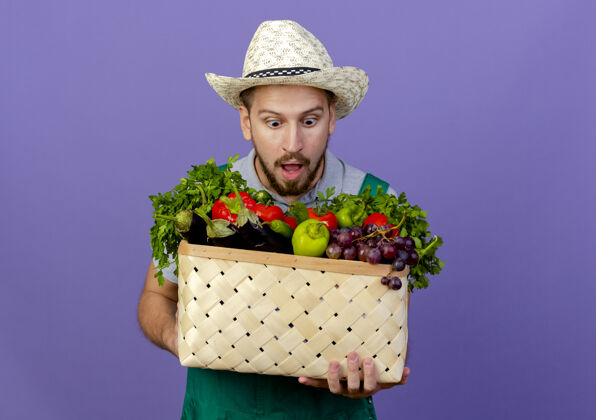 抱着年轻帅气的斯拉夫园丁穿着制服 戴着帽子 看着篮子里孤立的蔬菜帅哥帽子小伙子