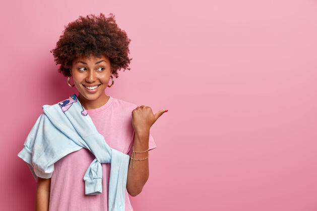 指向有着非洲头发的漂亮女孩帮着挑选最佳的选择 把拇指放在复印空间旁 做产品广告 开心地笑着 穿着玫瑰色的t恤和毛衣 肩上系着你的宣传片在这里毛衣显示乐观