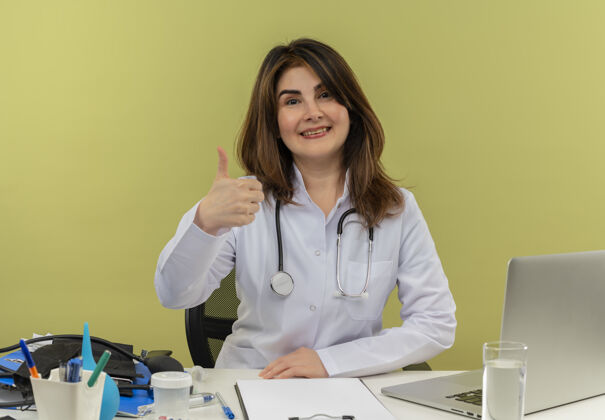 拇指微笑的中年女医生穿着医用长袍和听诊器坐在办公桌旁 拿着医疗工具剪贴板和笔记本电脑向上伸出大拇指个人剪贴板医生