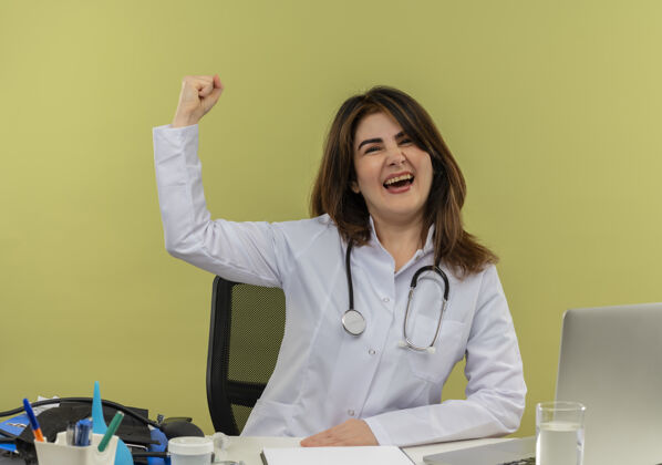 办公桌快乐的中年女医生穿着医用长袍和听诊器坐在办公桌前 拿着医疗工具剪贴板和笔记本电脑做“是”的手势笔记本电脑绿色欢乐