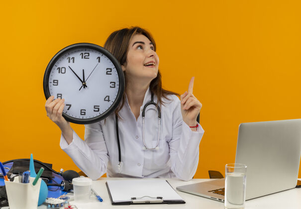 印象印象深刻的中年女医生穿着医用长袍和听诊器坐在办公桌前 拿着医疗工具剪贴板和笔记本电脑拿着时钟看着一边指向上工具长袍坐着