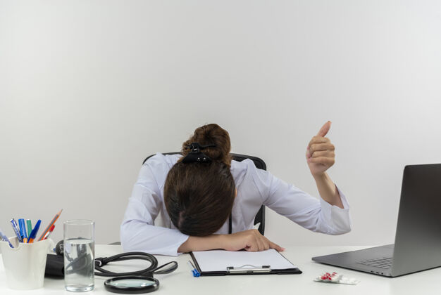 女性疲惫的年轻女医生穿着医用长袍和听诊器坐在办公桌旁 手持医疗工具和笔记本电脑 头放在办公桌上 大拇指向上竖起向上穿工具