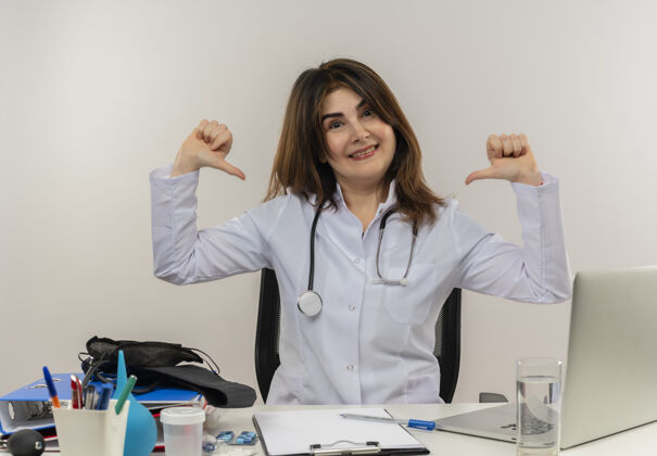 工具快乐的中年女医生穿着医用长袍和听诊器坐在办公桌旁 用医疗工具剪贴板和笔记本电脑指着自己听诊器指尖女性
