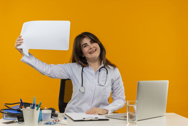女快乐的中年女医生穿着医用长袍和听诊器坐在办公桌旁 拿着医疗工具剪贴板和笔记本电脑 拿着聊天泡泡桌子橙色听诊器