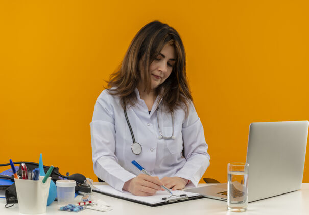 穿着专注的中年女医生穿着医用长袍和听诊器坐在办公桌旁拿着医疗工具笔记本电脑用笔在剪贴板上写字中年专注笔记本电脑
