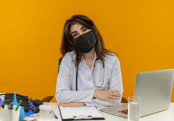 坐着中年女医生穿着医用长袍 听诊器坐在办公桌旁 医疗工具笔记本电脑和剪贴板被隔离笔记本电脑女橙色