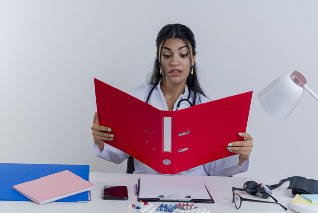 坐着印象深刻的年轻女医生穿着医用长袍和听诊器坐在办公桌前拿着医疗工具 看着文件夹抱着年轻人长袍
