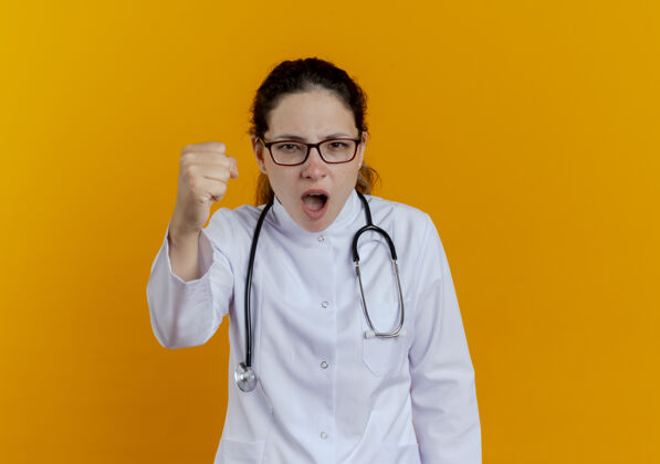 女士年轻严格的女医生 穿着医用长袍 戴着听诊器 戴着眼镜 伸出拳头医生穿年轻