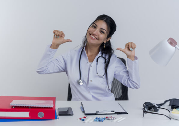 听诊器面带微笑的年轻女医生穿着医用长袍和听诊器坐在办公桌旁 拿着医疗工具指着自己孤立地看着自己指着工具年轻人