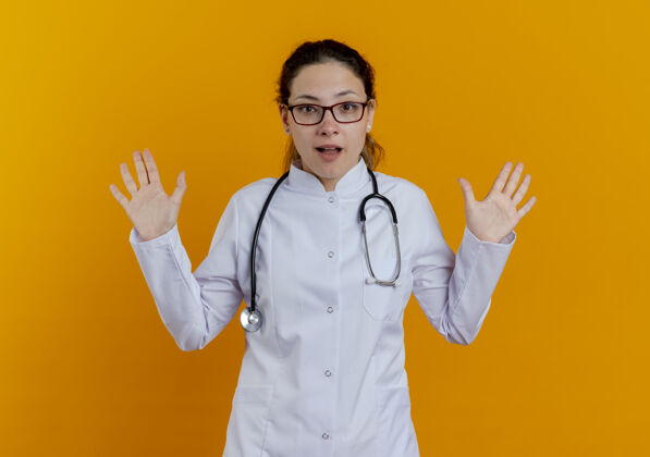 医生惊讶的年轻女医生穿着医用长袍和听诊器 戴着眼镜摊开双手橙色成年人女人