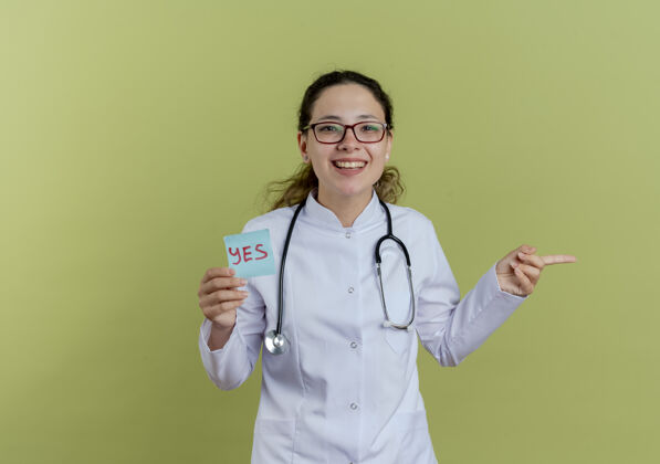 成人面带微笑的年轻女医生 穿着医用长袍 戴着听诊器 戴着眼镜 把纸上的笔记点隔离在一边听诊器医生女士