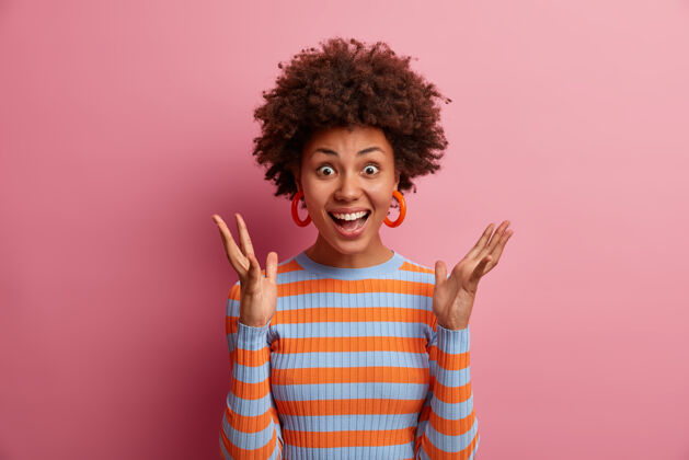 条纹兴奋快乐的非洲头发女人举起双手 听到可怕的消息 得到意想不到的惊喜 表达喜悦和惊奇 穿着休闲条纹毛衣 孤立在粉红色的墙上反应成人情绪