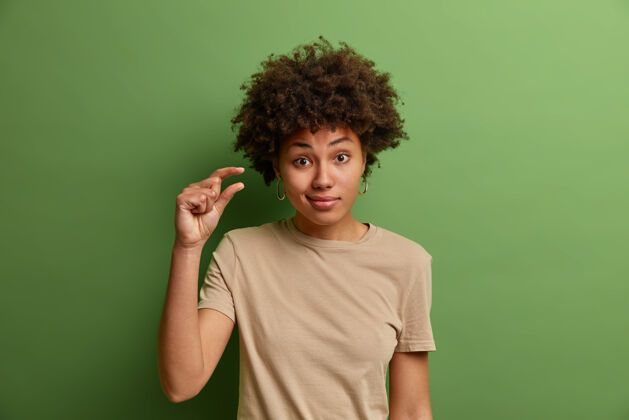 展示无动于衷的卷发女人表现出一些或多或少的姿态 显示几英寸或哨兵 少量或低比例的标志 穿着休闲服 隔离在绿色的墙上室内小比例