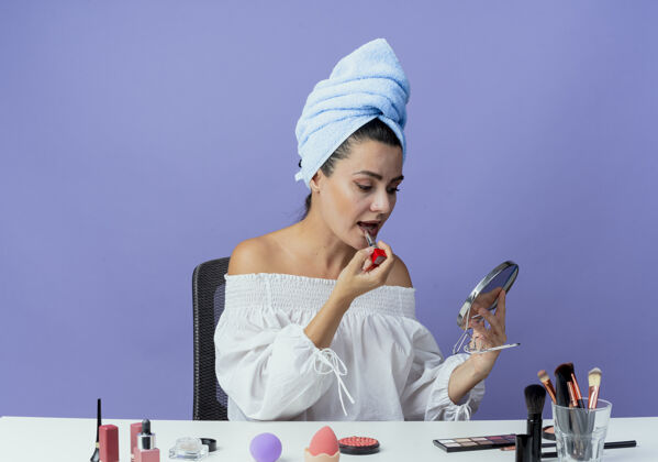 毛巾自信漂亮的女孩裹着发巾坐在桌子上 拿着化妆工具 拿着唇膏 看着镜子在紫墙上孤立紫色漂亮女人