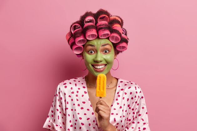 睡袍积极的女人接受美容程序 吃黄色的冰棒 戴卷发器做完美的发型 享受美味的冷甜点 穿睡衣 摆出粉红色的姿势请滚轴奶制品