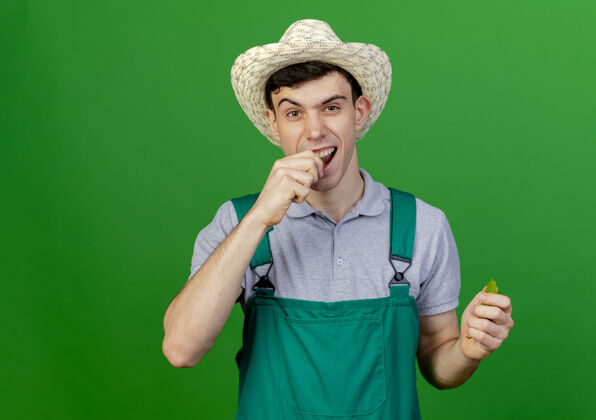 帽子快乐的年轻男园丁戴着园艺帽假装咬碎辣椒隔离在绿色背景与复制空间咬空间胡椒