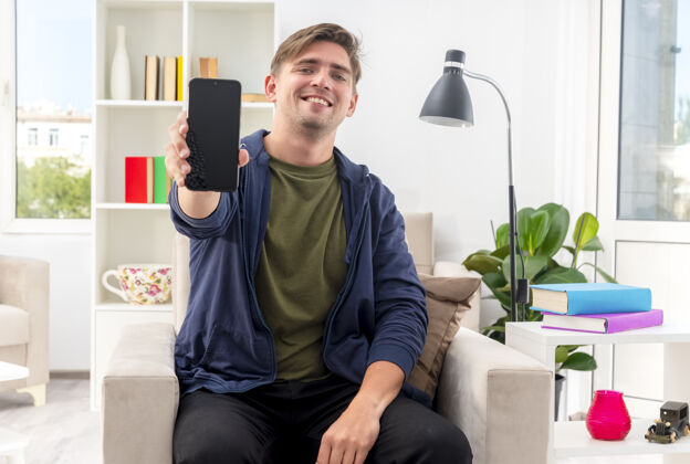微笑微笑的年轻金发帅哥坐在扶手椅上拿着手机看着客厅里的相机电话客厅抱着