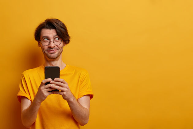 联系开朗的时髦小伙手持智能手机向朋友发送聚会上的搞笑照片 在网络中打字浏览 穿着随意 对着黄色墙壁摆姿势 复制空间使用姿势在线