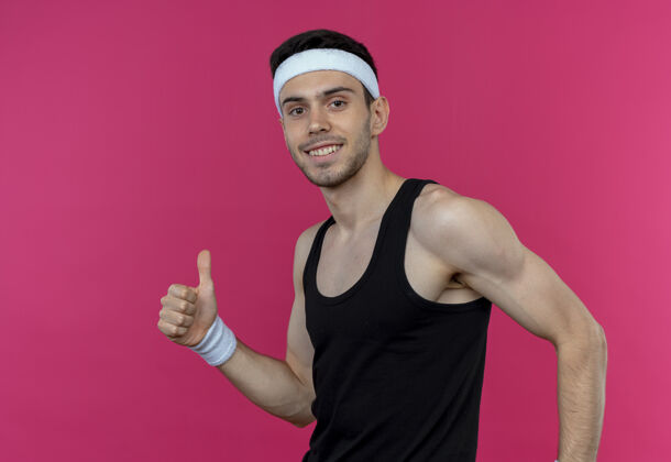 微笑戴着头巾的年轻运动型男子看着镜头 微笑着 高兴而积极地竖起大拇指站在粉色背景上拇指年轻运动