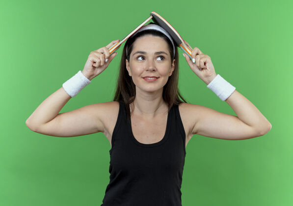 抱戴着头巾的年轻健身女士头上举着两个乒乓球拍 站在绿色背景上微笑着头带健身微笑