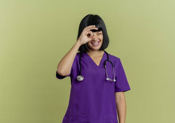 通过快乐的年轻黑发女医生穿着制服 手持听诊器 通过橄榄绿背景上的手指观察 并留有复印空间医生制服深色