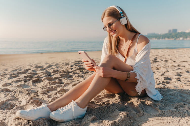 旅行者时尚年轻迷人的金发微笑的女人坐在沙滩上骑着自行车戴着耳机听音乐活跃服装休闲