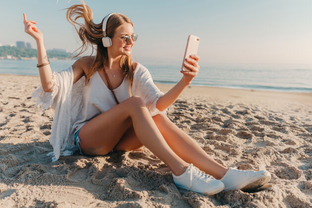 阳光年轻迷人的金发微笑女子在度假时坐在沙滩上打电话自拍沙滩音乐旅程
