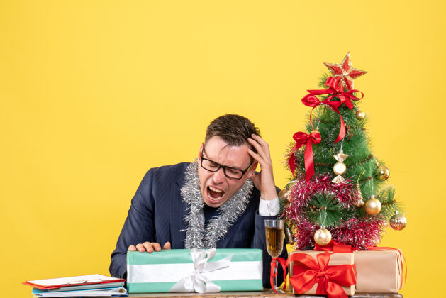 背景前视图愤怒的男人坐在圣诞树旁边的桌子上 呈现黄色背景女商人坐着主任