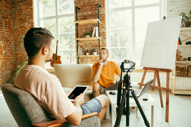 房子两位年轻的白人男性博主穿着休闲服 在家里用专业设备或摄像机录制视频采访博客 视频博客 视频博客在室内直播时聊天媒体男性记录