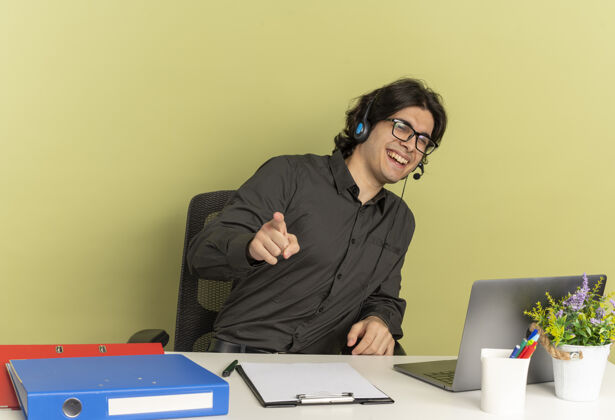 年轻年轻微笑的上班族曼农戴着眼镜戴着耳机坐在办公桌旁 拿着办公工具 用笔记本电脑指着隔离在绿色背景上的摄像头 还有复印空间使用光学坐着