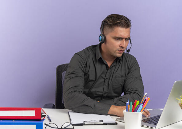 自信年轻自信的金发上班族戴着耳机坐在办公桌旁 手里拿着办公工具 看着孤立在紫色背景下的笔记本电脑 还有复印空间工具办公室耳机