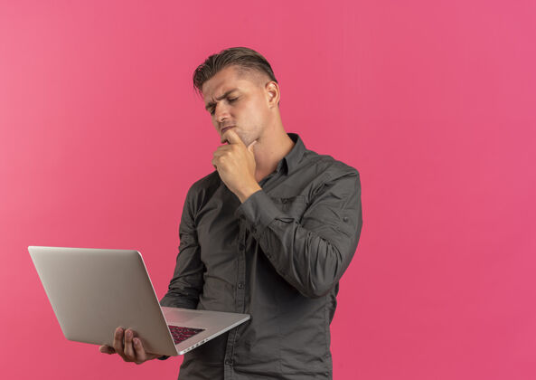 金发年轻迷茫的金发帅哥把手放在下巴上看着笔记本电脑孤立在粉红色的背景与复印空间困惑笔记本电脑英俊