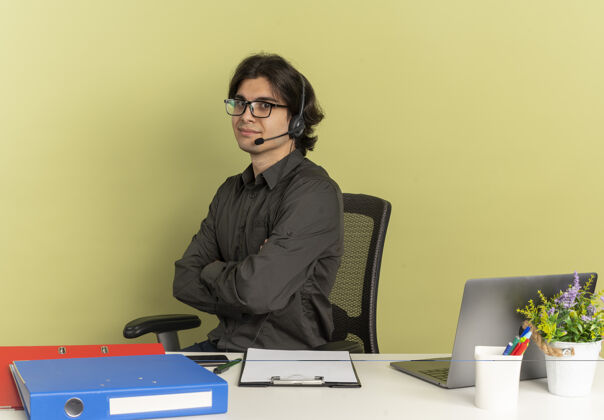 侧着年轻快乐的上班族曼农戴着眼镜 戴着耳机坐在办公桌旁 办公工具用笔记本电脑 双臂交叉 绿色背景 留有复印空间办公桌光学坐着