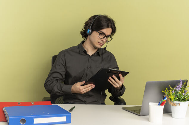 使用年轻自信的上班族戴着耳机 戴着眼镜 坐在办公桌旁 拿着办公工具 拿着笔记本电脑 看着隔离在绿色背景上的剪贴板 还有复印空间拿着工作人员眼镜