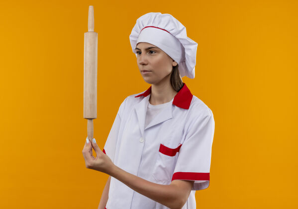 烹饪年轻自信的白人厨师女孩穿着厨师制服 拿着擀面杖 看着橙色背景上的复制空间举行厨师女孩