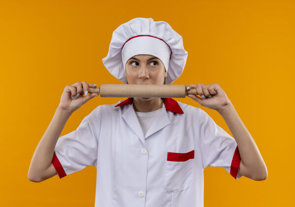 惊喜穿着厨师制服的年轻白人厨师女孩拿着并假装咬着橙色背景上的擀面杖 并留有复印空间厨师假装咬
