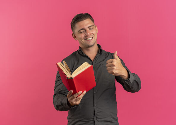 微笑年轻微笑的金发帅哥拿着书 竖起大拇指孤立地放在粉色背景上 留着复印空间金发抱着年轻