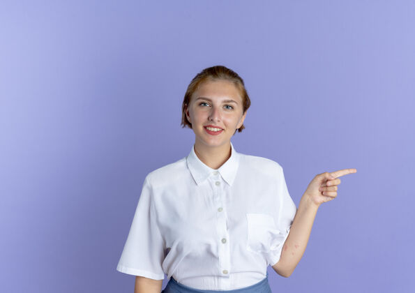 尖年轻的微笑金发俄罗斯女孩点在一边孤立的紫色背景与复制空间年轻女孩金发