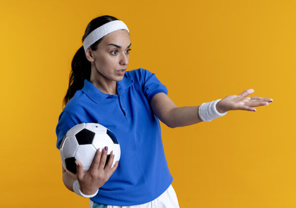 困惑年轻困惑的高加索运动女性戴着头带和腕带拿着球指着橙色背景上孤立的一边 并留有复制空间球侧线穿