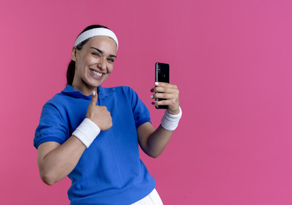 电话年轻微笑的白人运动女性戴着头带和腕带拇指向上拿着手机在粉红色背景上与复制空间隔离抱着戴运动