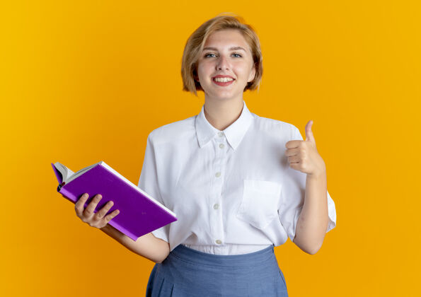金发年轻微笑的金发俄罗斯女孩拿着书 竖起大拇指孤立在橙色背景与复制空间抱着拇指女孩