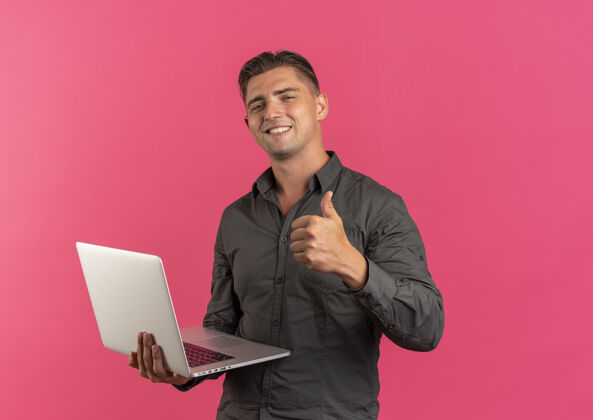 帅气年轻微笑的金发帅哥拿着笔记本电脑 竖起大拇指孤立地放在粉色背景上 留着复印空间笔记本电脑拇指金发
