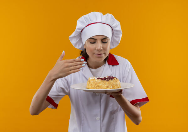 年轻身着厨师制服的年轻高加索厨师女孩高兴地拿着盘子 假装闻到蛋糕的味道 盘子被隔离在橙色的背景上 并留有复印空间制服女孩厨师