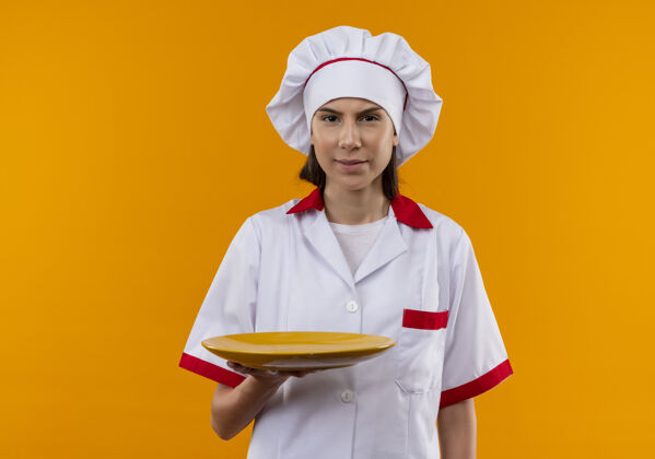 相机穿着厨师制服的年轻恼怒的白人厨师女孩拿着盘子 看着隔离在橙色背景上的相机 还有复印空间厨师女孩烹饪
