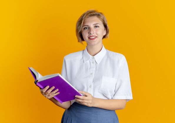 相机年轻的微笑的金发俄罗斯女孩拿着书看着相机孤立在橙色背景与复制空间微笑拿着金发